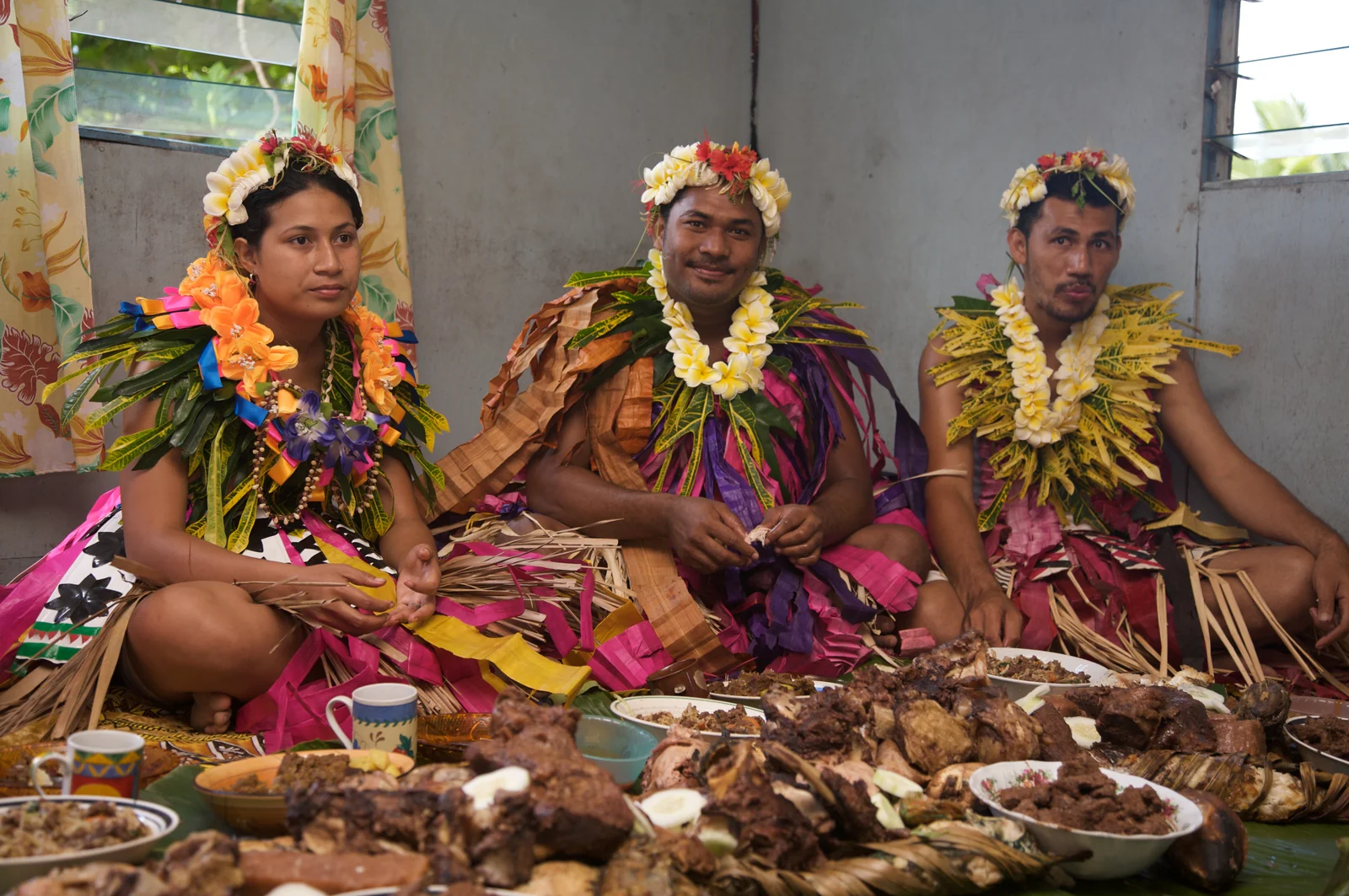 Three people in flower garlands seated behind dishes at a Fijian feast