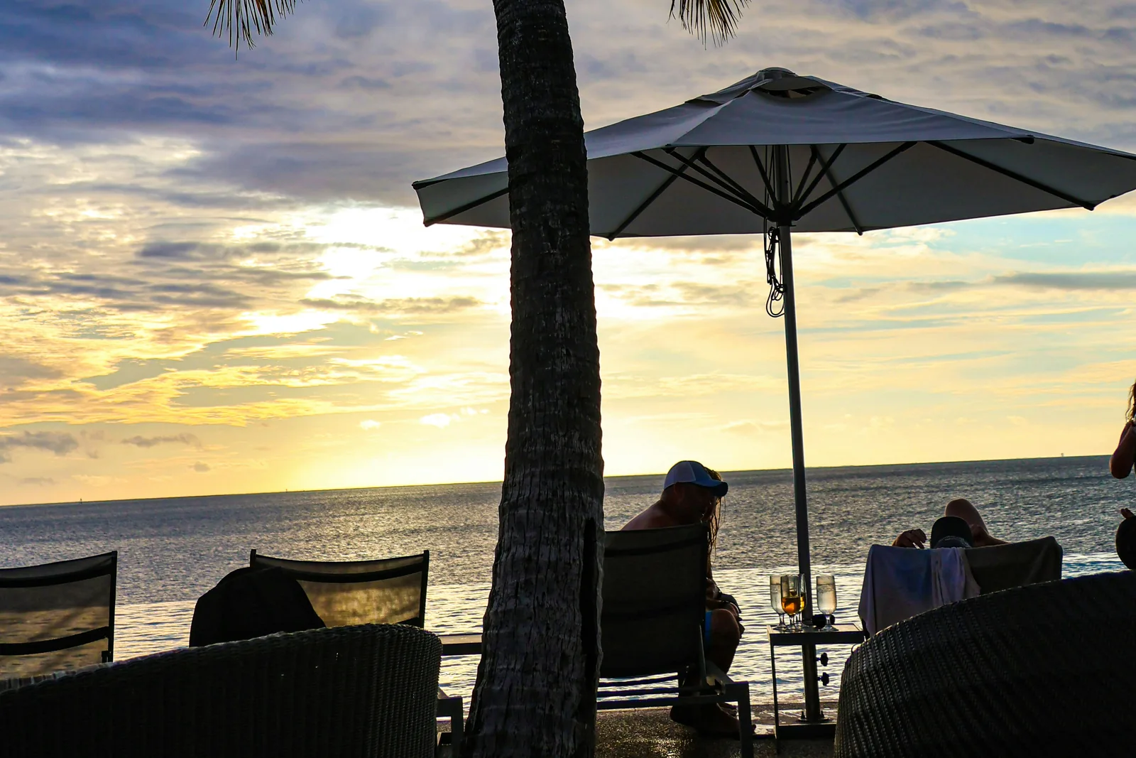 Guests with drinks beside the ocean under a palm tree and umbrella at sunset in Momi Bay, Fiji.